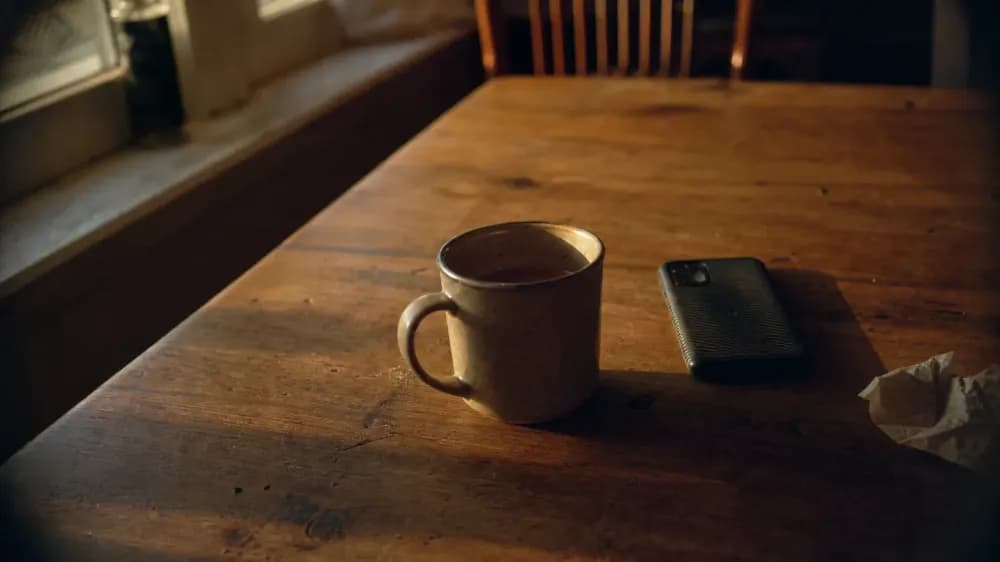 A stoneware mug of tea left untouched on a scratched wooden table beside a phone lying face down, in weak evening light from a window at the left, with nobody in the room.
