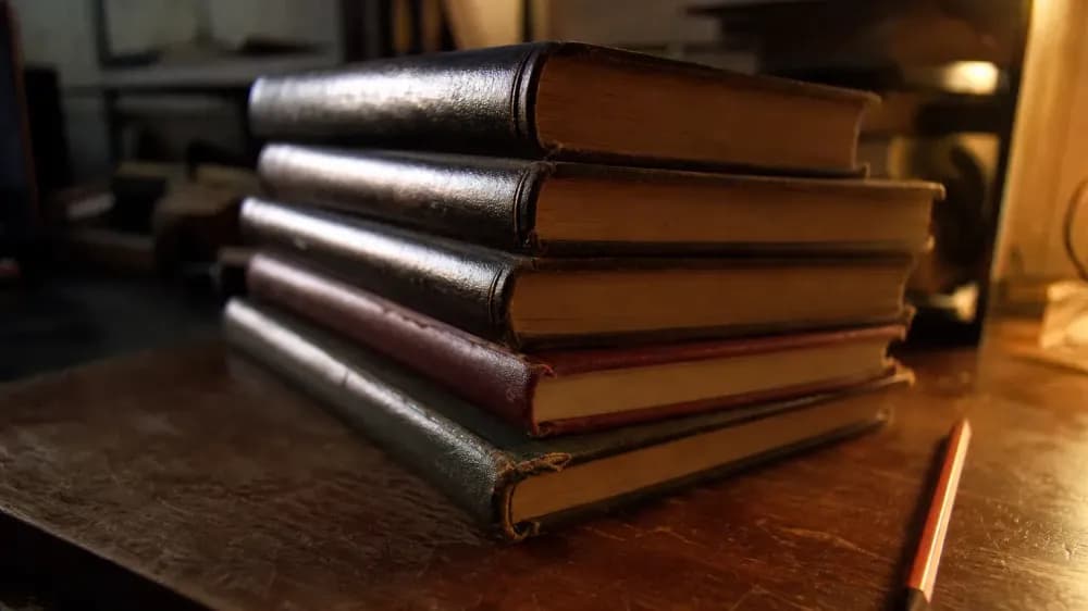 A stack of worn hardback journal volumes on a desk under a low lamp, spines angled away from the camera so no title can be read.