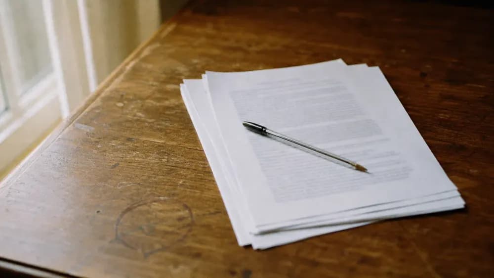 A loose stack of printed report pages on a worn wooden desk beside a window, a ballpoint pen lying across the top page, photographed at a low angle so the printed type reads only as soft grey lines.