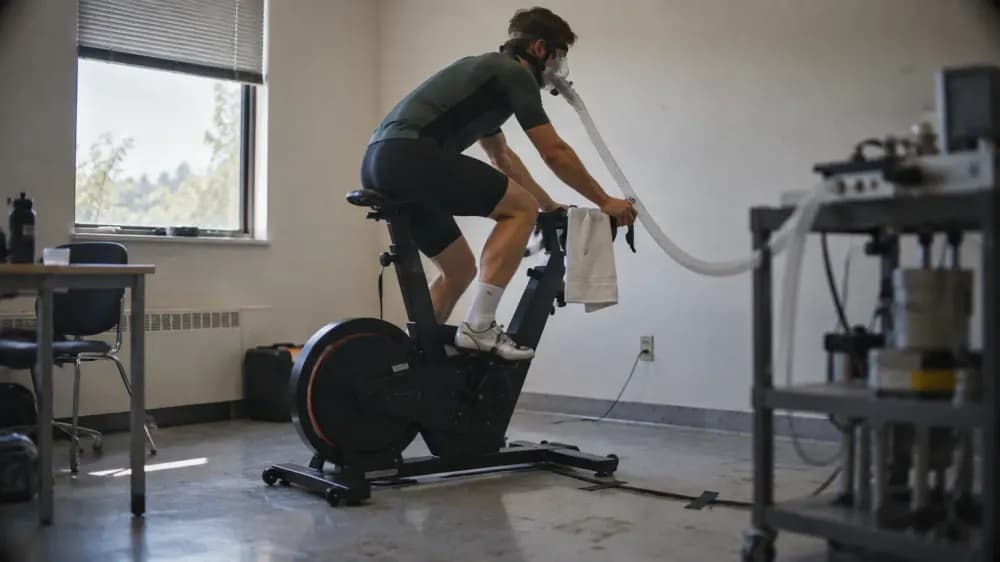A cyclist on a stationary lab bike wearing a breathing mask connected to a gas-analysis hose, photographed from the side in a plain exercise physiology lab.