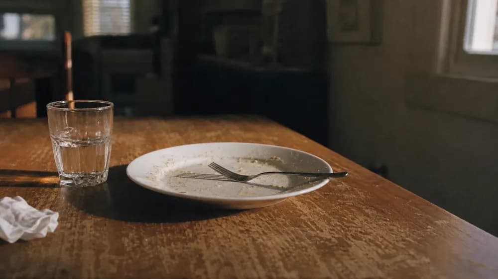 A used dinner plate and fork left on a scuffed wooden table in low evening light from a single window, with a half-empty water glass beside them and nobody in the room.