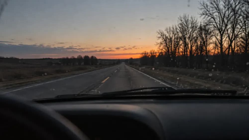 An empty two-lane road at dusk seen through a car windshield, wipers at rest, the dashboard a dark silhouette against a pale sky.