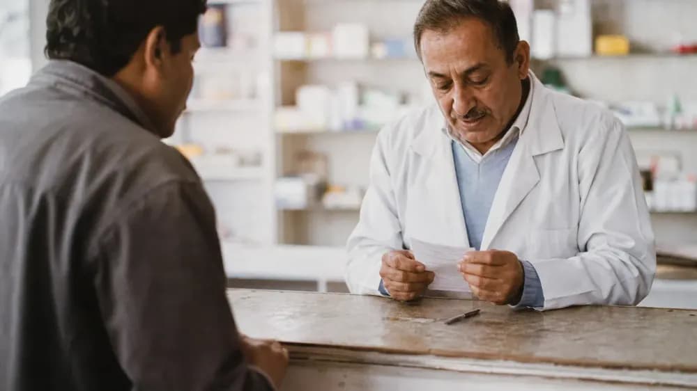 A person at a pharmacy counter handing a handwritten list of everything they take across to a pharmacist.