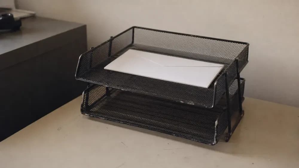 Two stacked wire desk trays on a plain office desk, the upper tray holding a single unmarked white envelope and the lower tray empty.