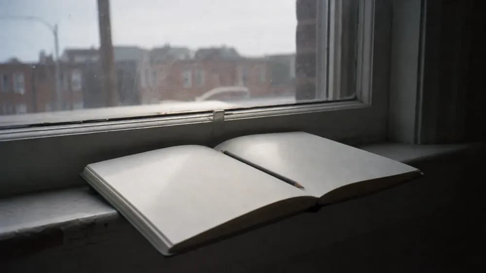 An open notebook with a pencil resting in the gutter, left on a wide windowsill in flat overcast daylight, with the page angled away so no writing is legible and nobody in the room.