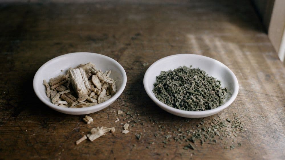 Two small plain white dishes side by side on a worn wooden bench, one holding pale woody root pieces, the other holding dried crumbled green leaf.