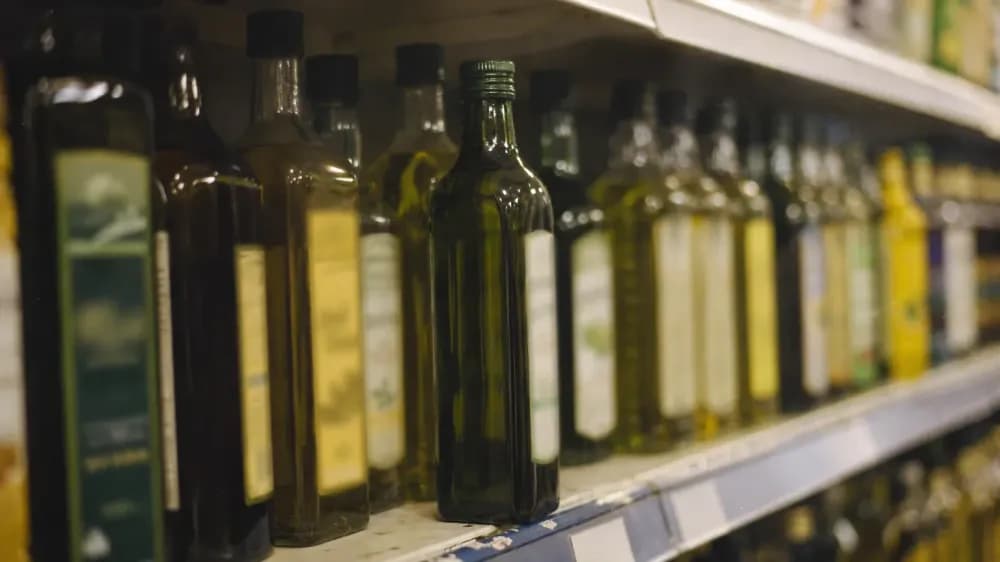 A grocery shelf of cooking oil bottles seen slightly from the side in ordinary store lighting, one dark green bottle pulled forward from the row, labels turned away and unreadable.