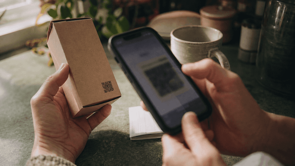 A person holding a plain unbranded cardboard tincture box in one hand and a phone in the other, scanning the code square printed on the box.