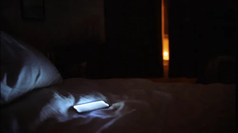 A dark bedroom at night with the glow of a phone screen lighting a rumpled duvet, the screen content out of focus and unreadable.