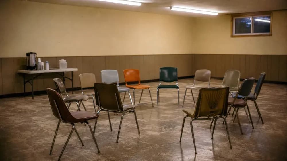 An empty community meeting room in the evening, stacking chairs arranged in a loose circle, a coffee urn on a folding table against the wall.